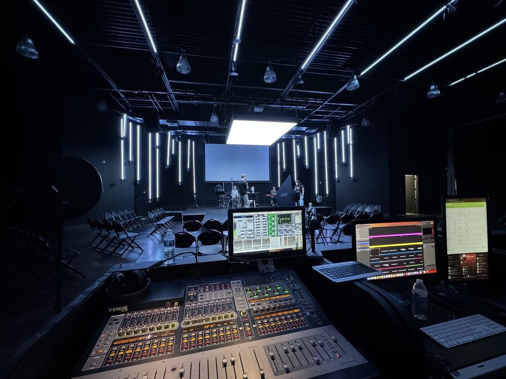 Front-of-house mixing console facing a black-box stage with hanging light fixtures