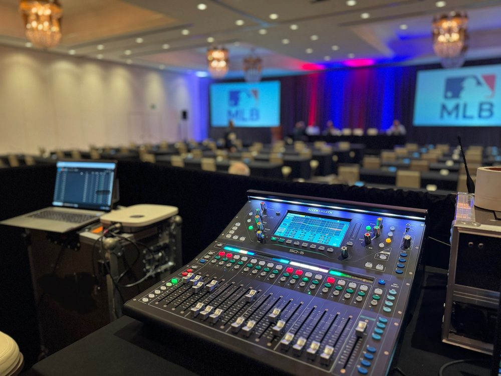 Front-of-house audio mixing console with MLB branded screens on stage in the background